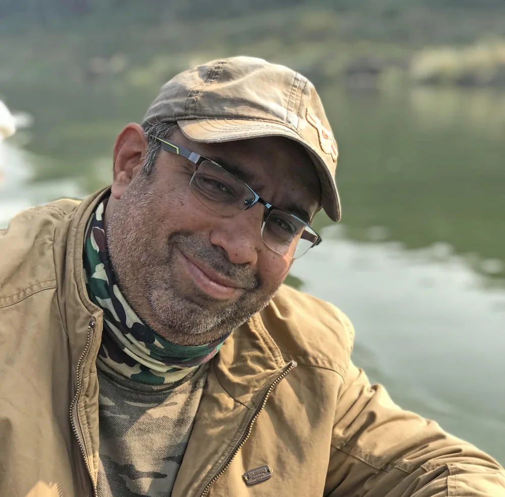 Wildlife photographer Vinit Arora smiles in a cap and safari jacket beside a calm river in the field.